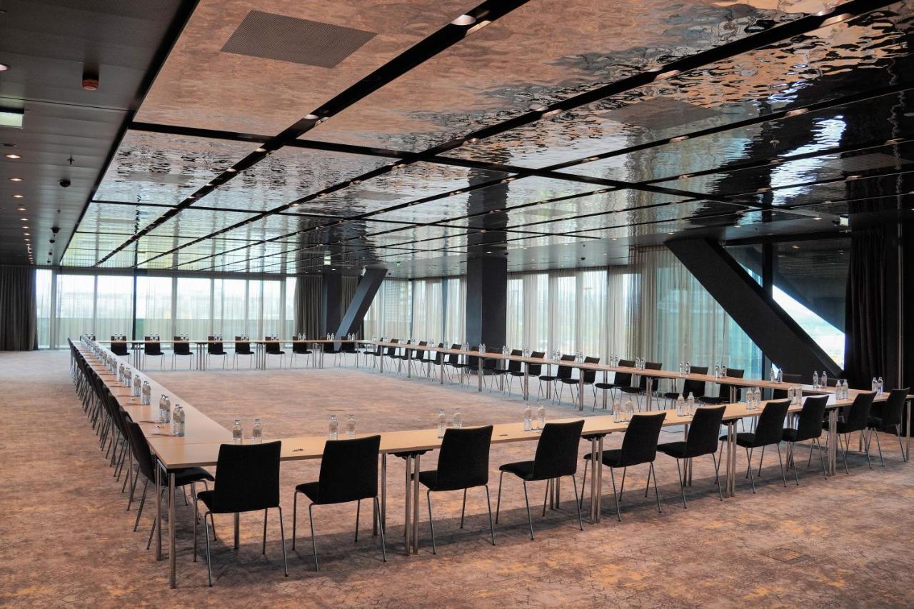 Hotel Melia Vienna Esterno foto The photo shows a modern conference room with a long rectangular table arranged in a U-shape. The table is flanked by multiple black chairs. The room features a sleek, reflective ceiling that gives a contemporary feel, and large windows allow natural