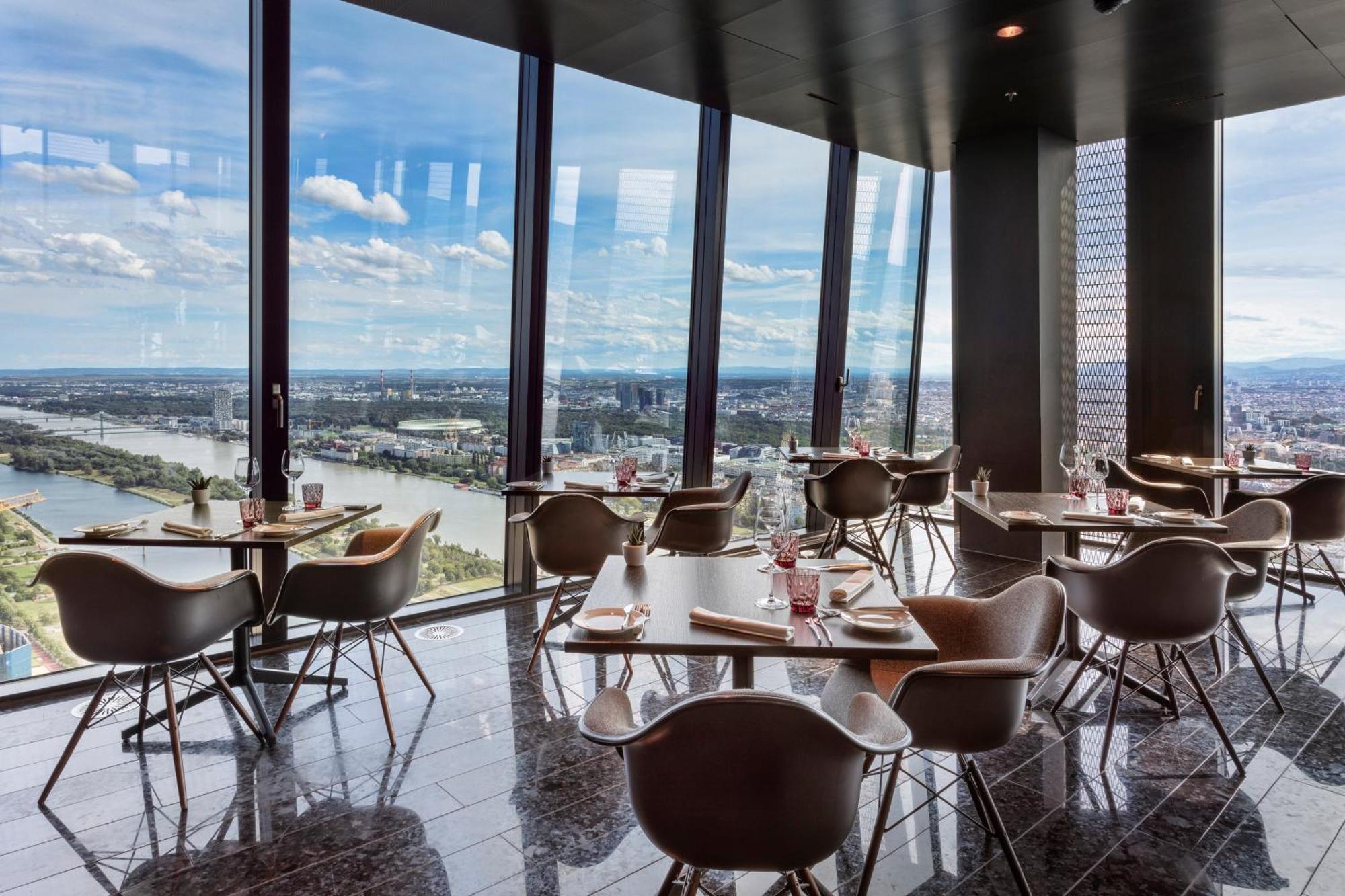 Hotel Melia Vienna Esterno foto The photo shows a modern dining space with a sleek design, featuring several tables and chairs arranged near large floor-to-ceiling windows. These windows provide a panoramic view of a river and surrounding landscape. The dining area has a contempora