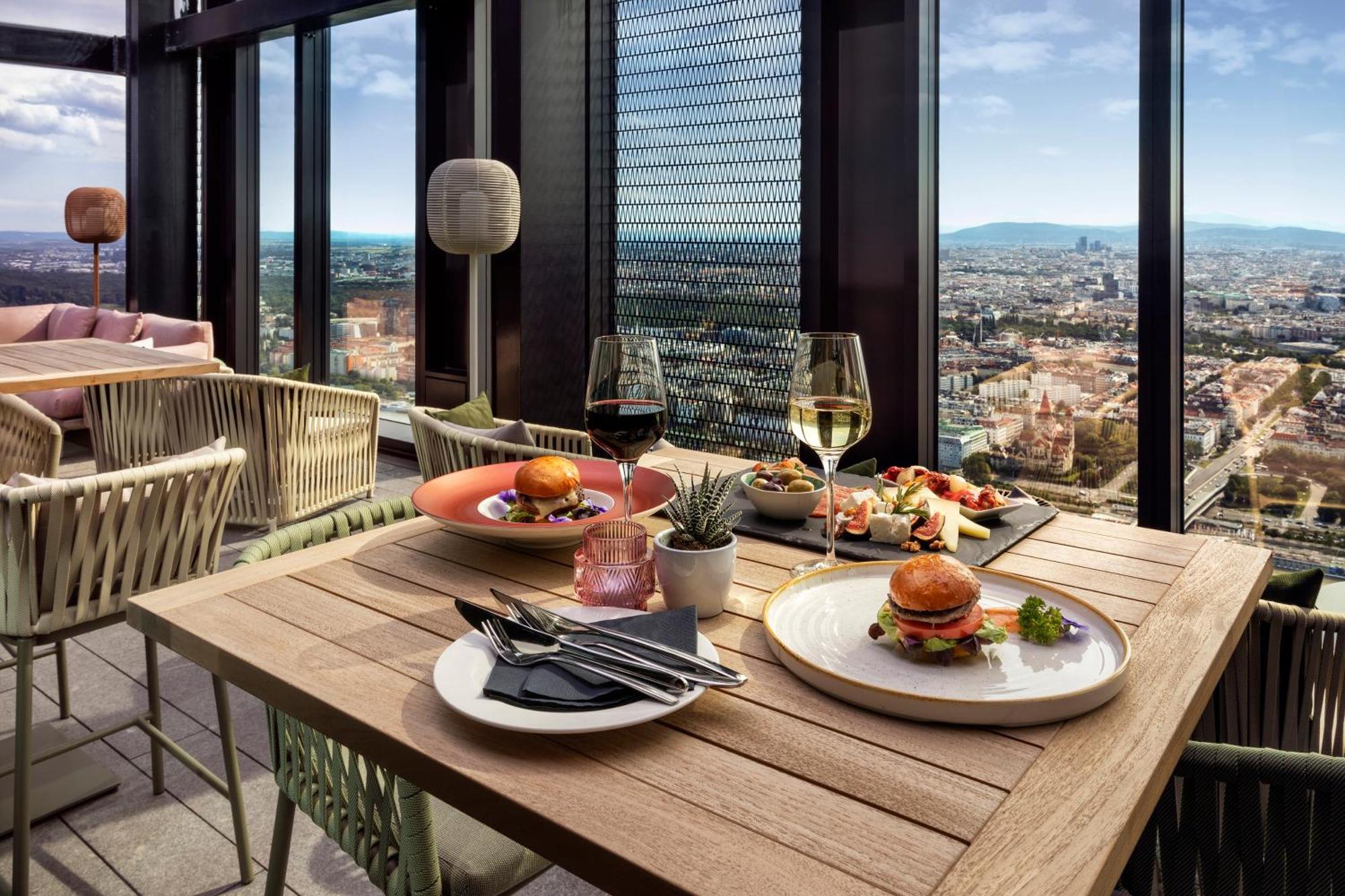 Hotel Melia Vienna Esterno foto The photo shows a stylish dining setup on a high rooftop or terrace, with a panoramic view of a city skyline in the background. 

There are two tables made of wood; one features two glasses of white wine and an assortment of dishes, including a plate