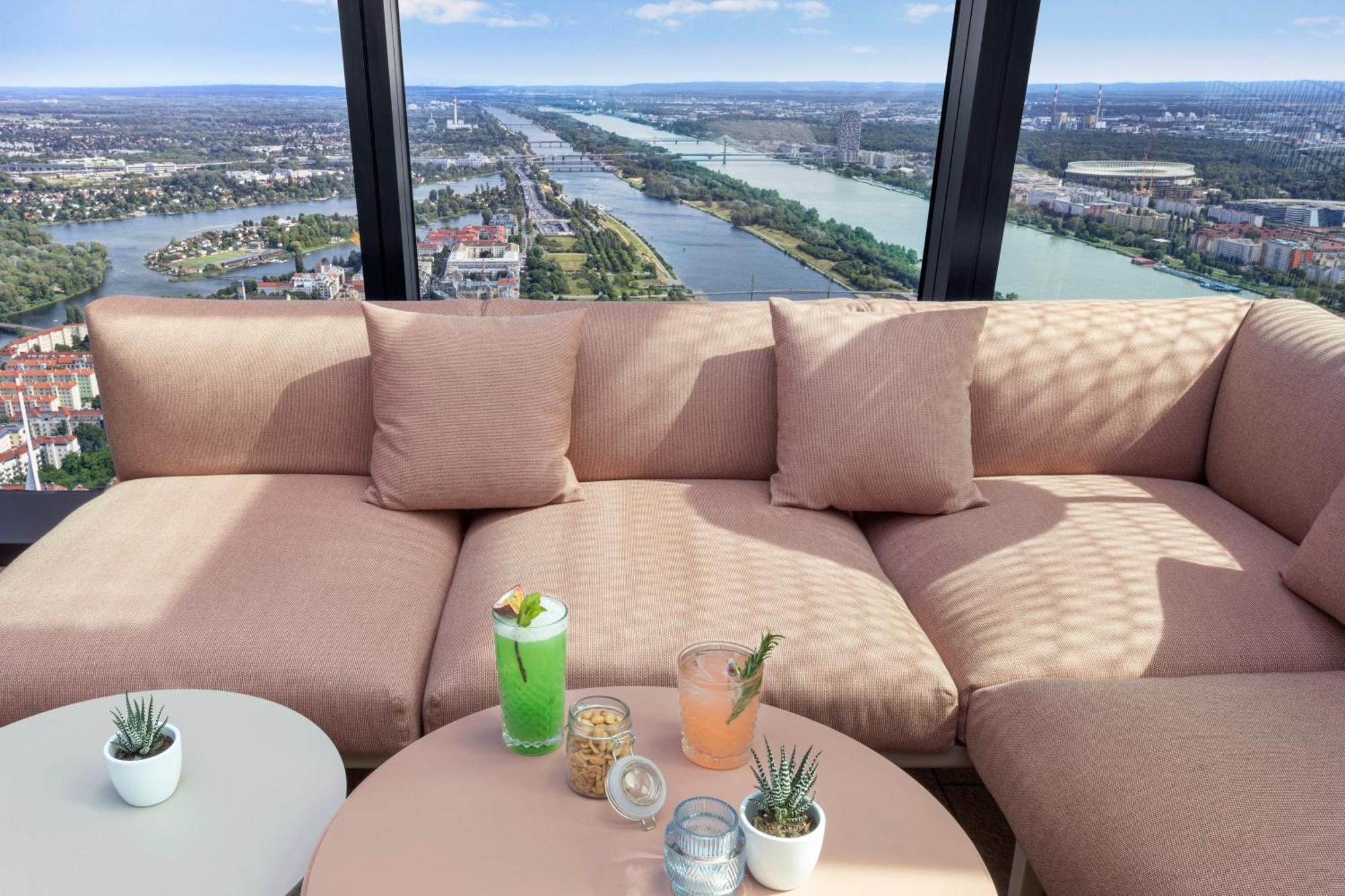 Hotel Melia Vienna Esterno foto The photo shows a cozy seating area with a large, soft, light pink couch and some small decorative pillows. In front of the couch, there is a round table with a few drinks placed on it: a green cocktail with a garnish, a light pink drink, and some sn