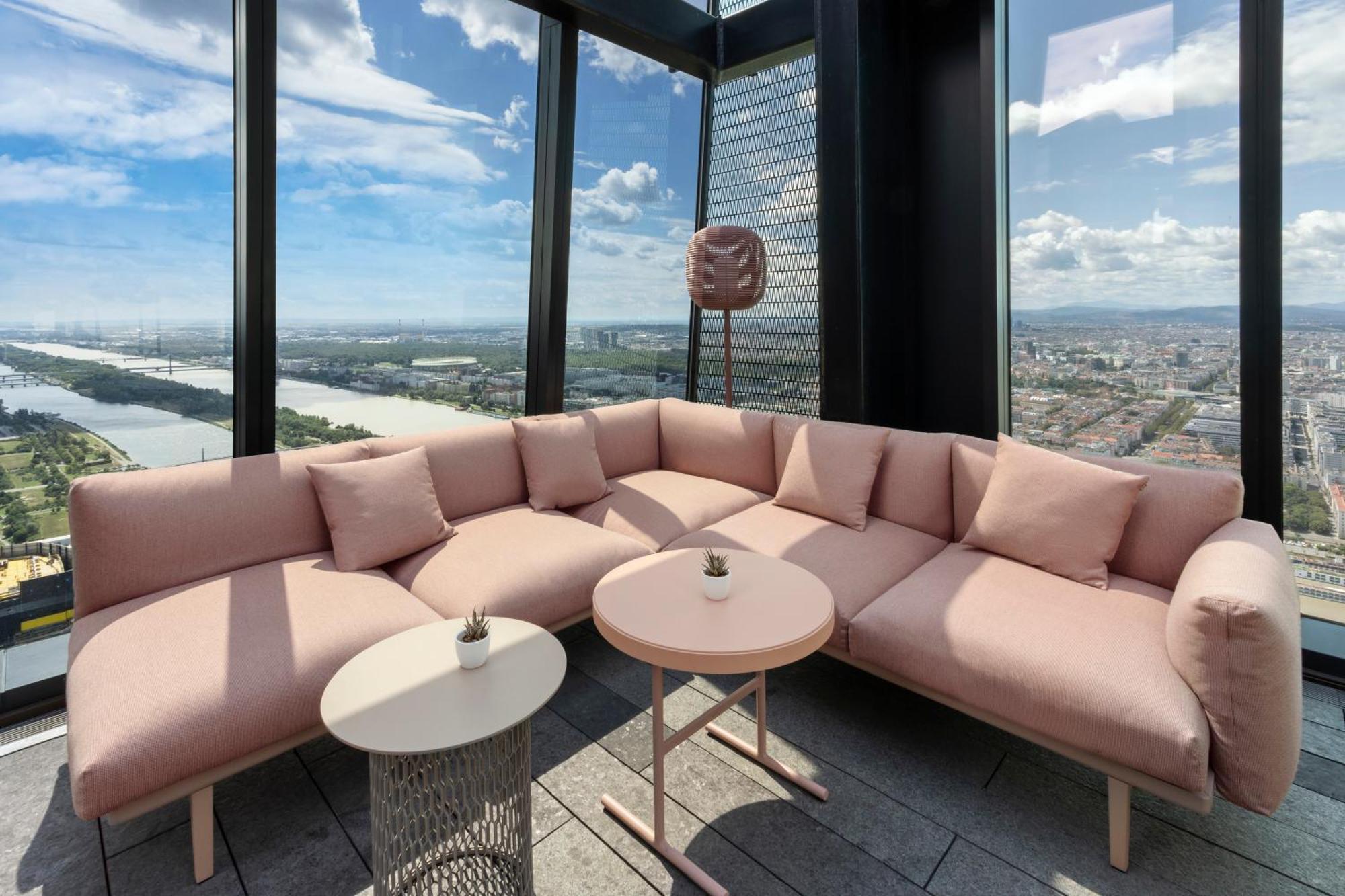 Hotel Melia Vienna Esterno foto The photo shows a modern lounge area with a large, L-shaped pink couch adorned with several soft cushions. In front of the couch, there are two tables: one round and one with a more intricate design. The space is located in a high-rise building, offe