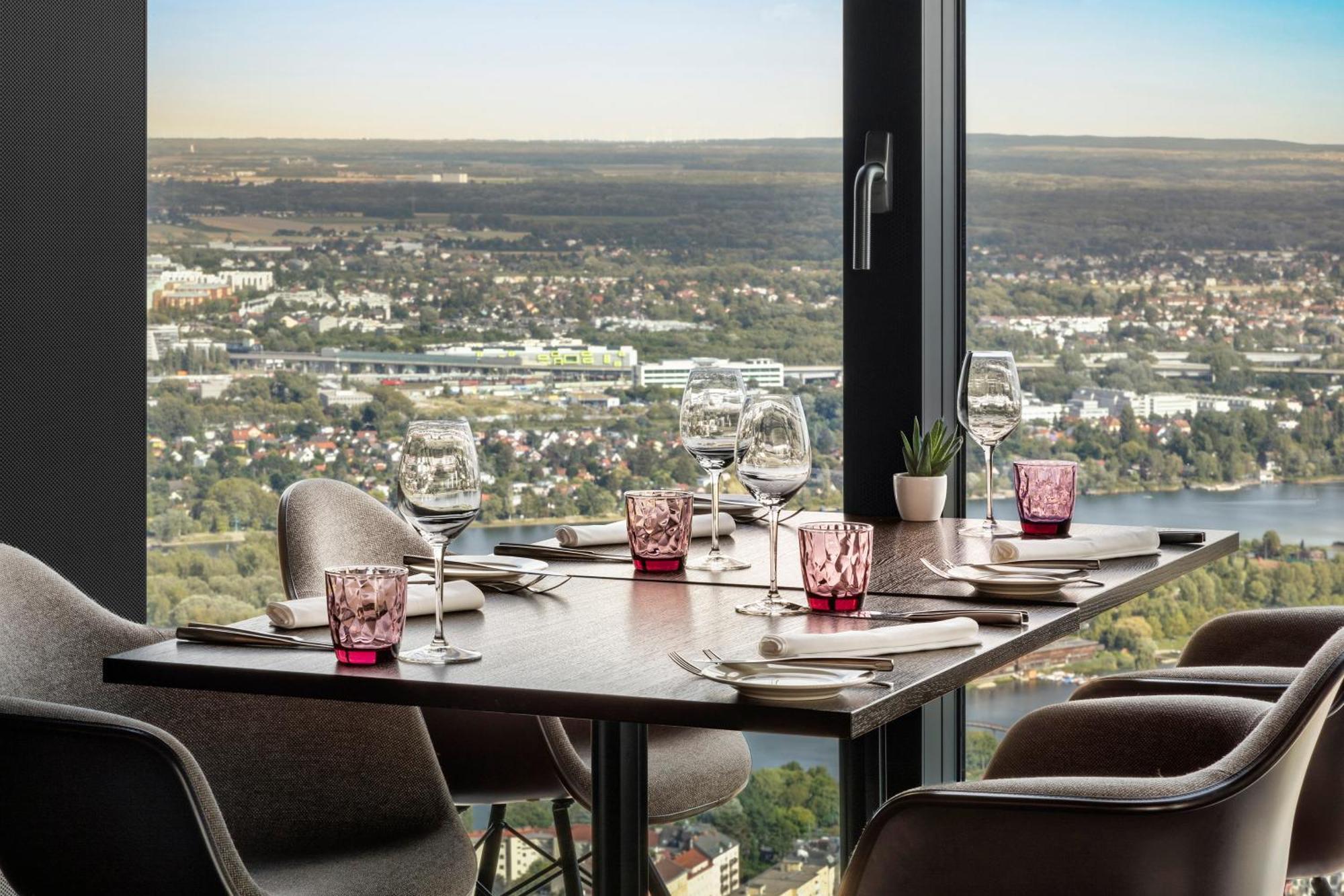 Hotel Melia Vienna Esterno foto The photo shows a dining table set up in a restaurant with a view. The table has dark wood and features several dining essentials arranged neatly. There are elegant wine glasses, pink-tinted glass holders, and plates set beside them. A small plant is