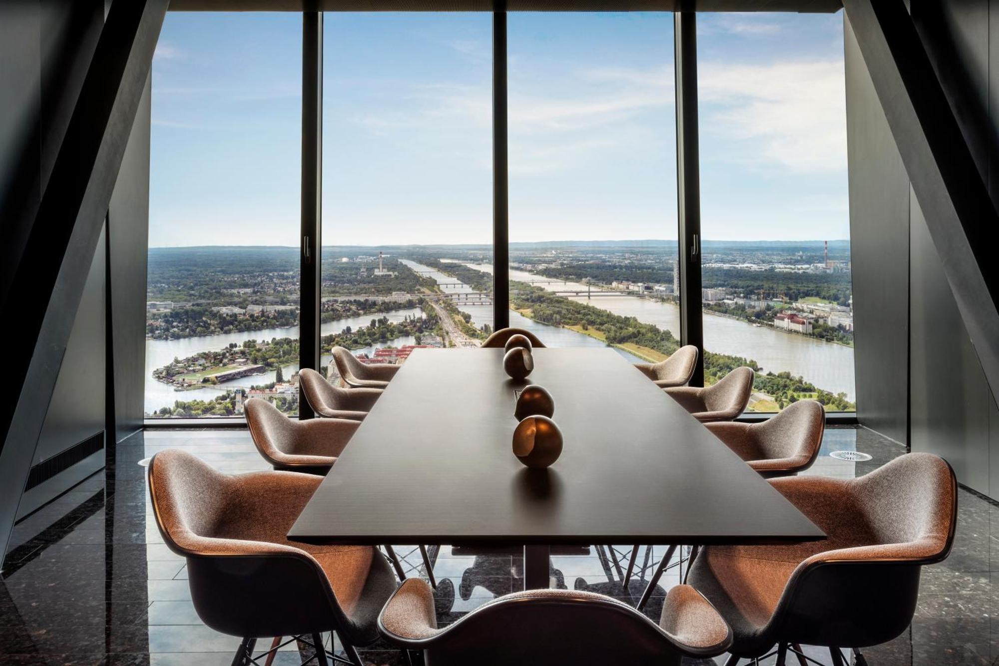 Hotel Melia Vienna Esterno foto The photo depicts a modern conference room with a long table surrounded by several stylish chairs. The room features large floor-to-ceiling windows that offer an expansive view of a landscape featuring water, greenery, and man-made structures in the 